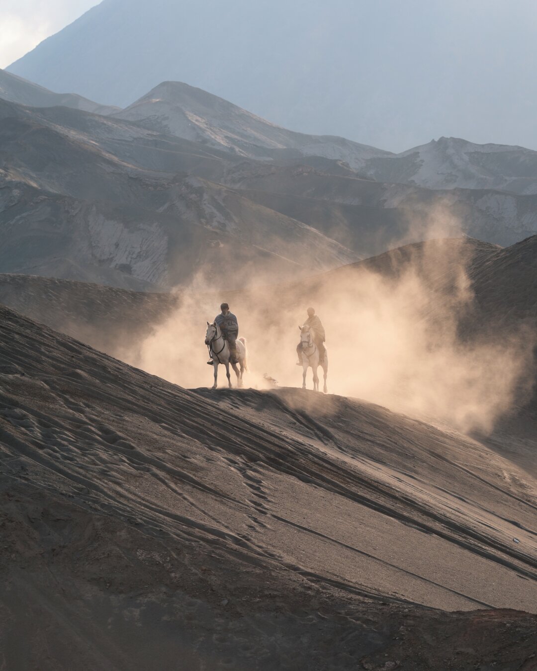 Two horsemen on mountainous volcanic plain trotting towards the camera, raising a lot of sun-list volcanic sand