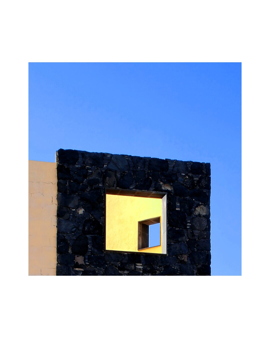 The image is in square format, showing a square-shaped facade of dark raw volcanic rocks against a clear blue sky. A square window opens to a piece of a yellow wall, also with a square window, framing a piece of the sky - creating a matrjoschka effect.