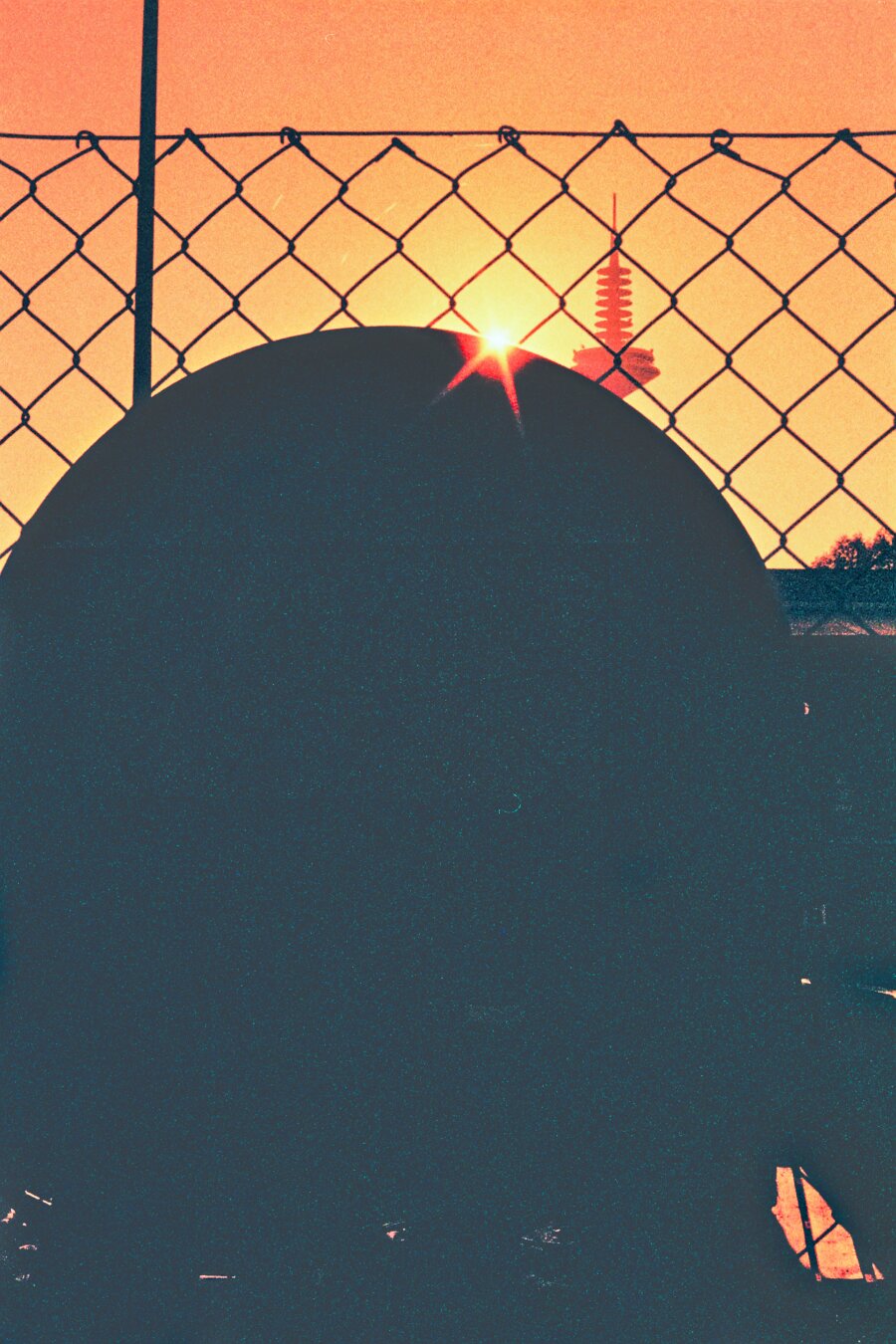 Analog color photo of the sun which is half covered by a black object in the foreground. Frankfurt television tower („Europaturm“) is next to the sun. The photo was taken through a wire mesh fence. Due to the film used the colors of the photo are red, orange and black.