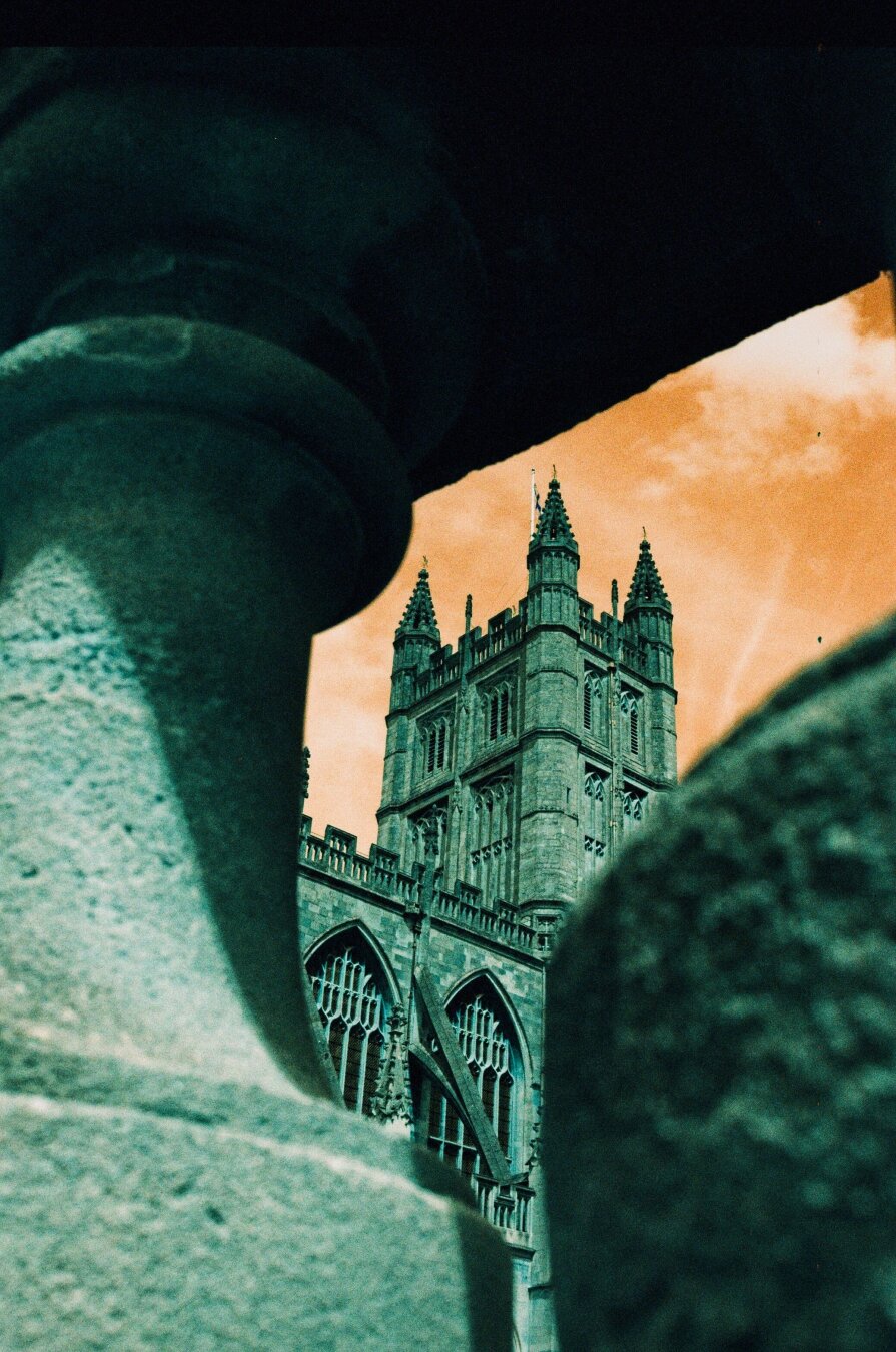 Analog color photo of a tower of the Abbey Church in Bath, GB. The tower is framed by parts of a balustrade in the foreground. Two large gothic windows can be seen from this perspective. Due to the turquoise film used the sky is orange and the stones green tinged.