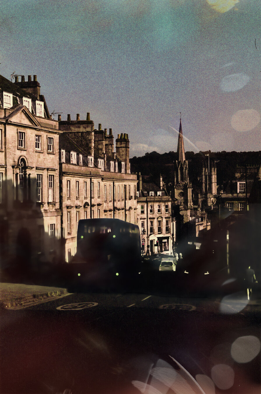 Analog color photo of a road in bath leading downwards. Apartment houses on the left, two busses on the street which are only seen as shadows, in the background, downwards the street is a church with a long tower spire. The photo is grainy and filled with plenty of glare spots. As the film used gives the photo a turquise touch and the original photo became way too dark, this shot has been edited in colors, brightness and glare spots.