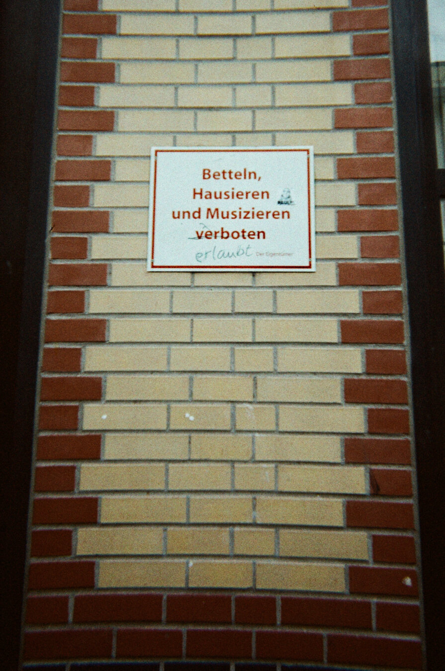 Analog color photo of a sign on a wall saying: "Betteln, Hausieren und Musizieren verboten" ("Begging, peddling, and busking prohibited"). One cheeky smart ass crossed out the word "verboten" ("prohibited") and added handwritten "erlaubt" ("allowed"). Not in the photo: The beggar, the vacuum cleaner salesman and the big band right in my back, your esteemed photographer.