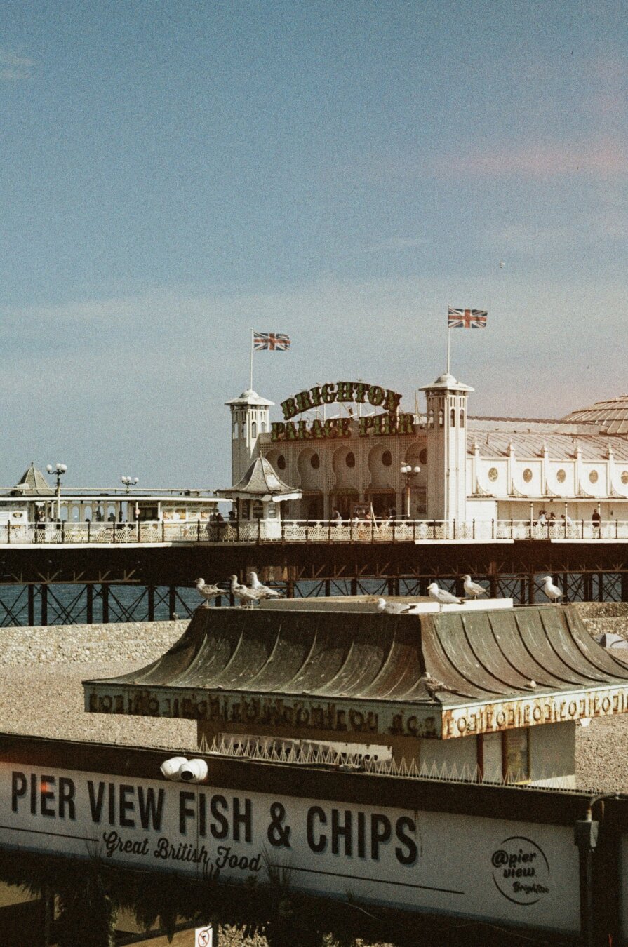 Analog color photo of the pier of Brighton, GB. In the foreground a sign on a biilding advertises „Pier view fish & chips“ (of course), some seagulls sittin on the roof of that building (probably waiting for their order to come). The „Brighton Palace Pier“ is in the background, two Union Jack flags on each side of the entrance.