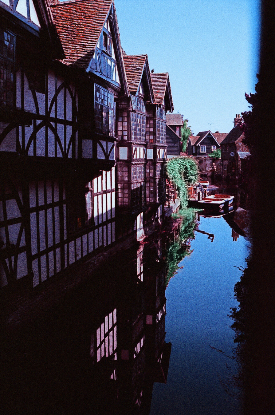 Analog color photo of three old frame houses standing side by side. The houses reflect in that small canal in front of the houses.