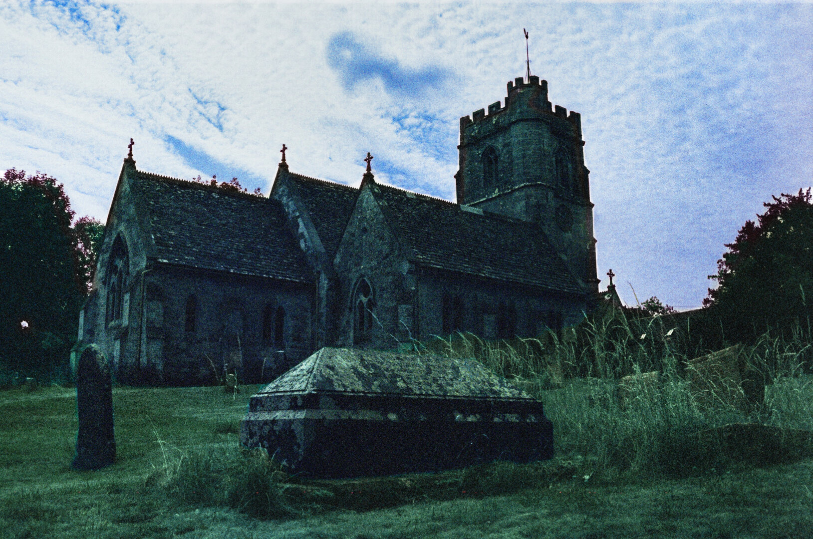 Analog color photo of a cemetary in Nympsfield in the Cotswolds, UK. A stone casket in the foreground, a gothic church in the background. Colors of the photo are edited, as the original shot has, due to the film used, a turquoise touch.