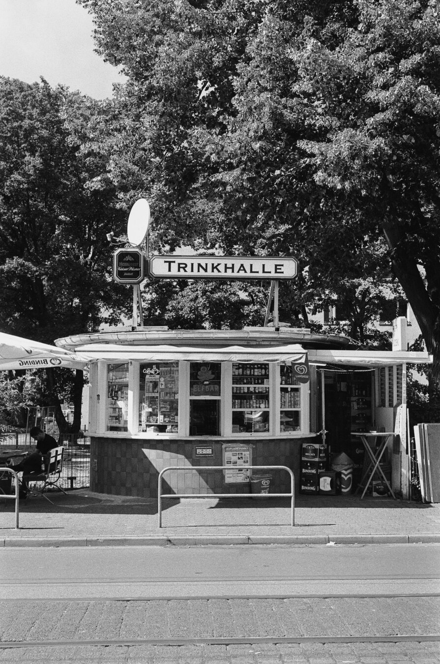 Analog black and white photo of a kiosk. Four shop windows with merchandise on display are visible; in the middle is an open window for sales. On the roof is a sign with the inscription „Trinkhalle“ (translated „drinking hall“, which totaly kills the spirit of that word). In Frankfurt they are called „Wasserhäuschen“ („little water house“).