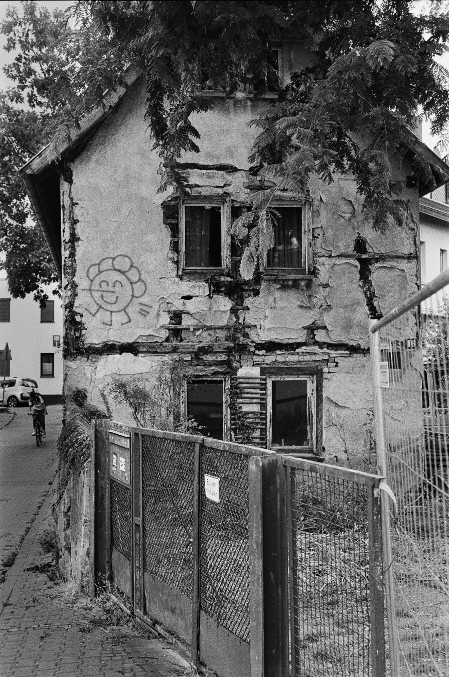 Analog black and white photo of an old, dilapidated house in Frankfurt-Ginnheim. Six windows on three floors are visible on the narrow side of the house. A fence is in front of the house. To the left of the building, a person is riding a bicycle.