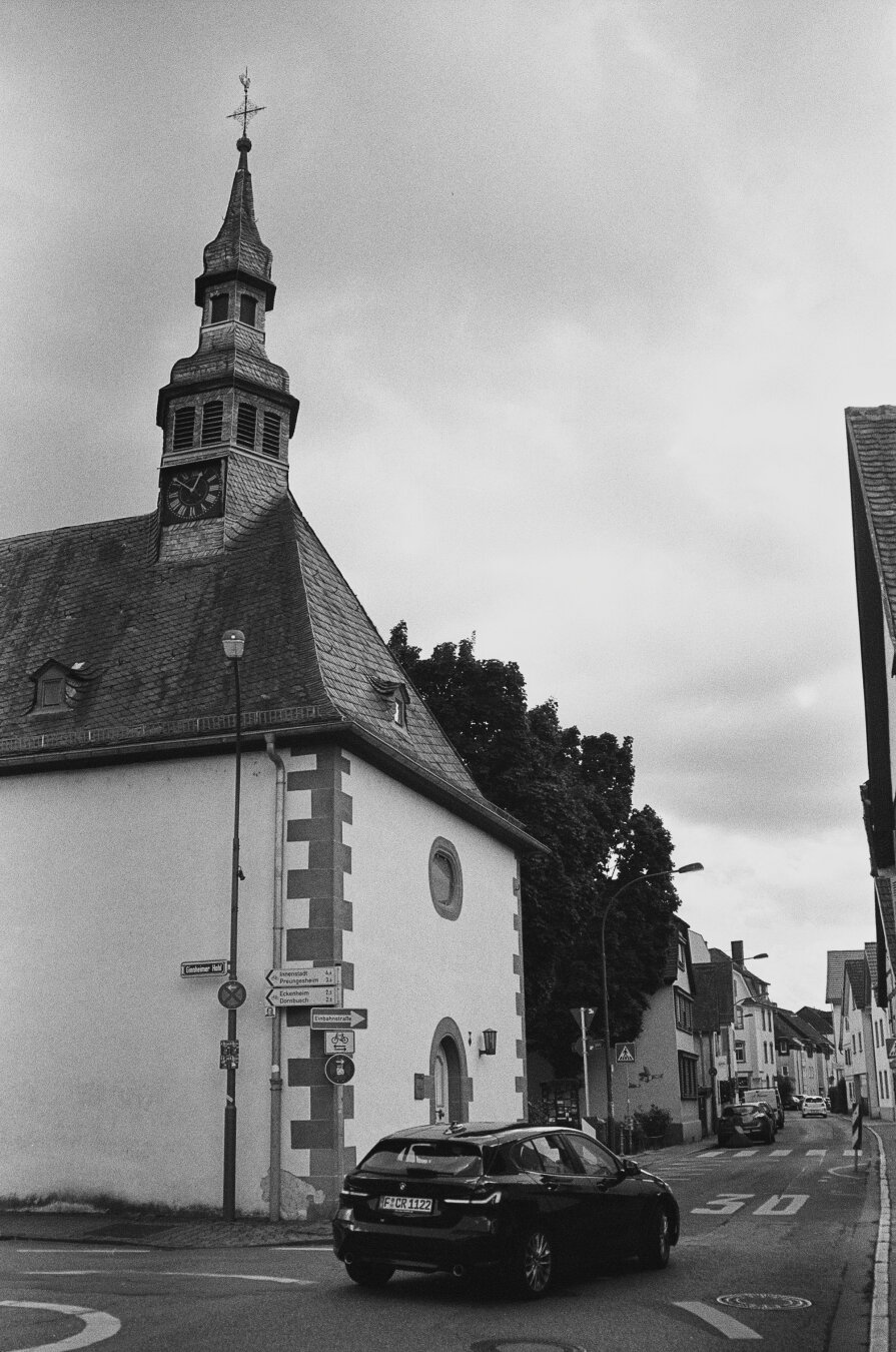 Analog black and white photo of the Bethlehem Church in Frankfurt Ginnheim. The building is white, the roof is black with a church steeple. The church is located on a street corner. A car turns into the street in front of the church.