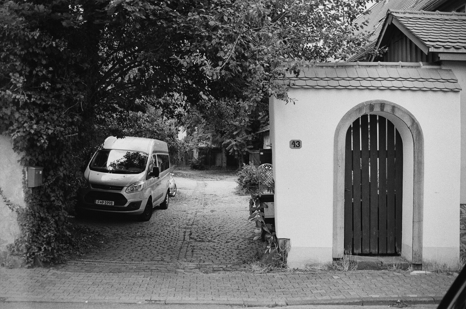 A former farm: A wide entrance to a courtyard with cobblestone. A wodden door the right side, a van parked under a tree on the left.
