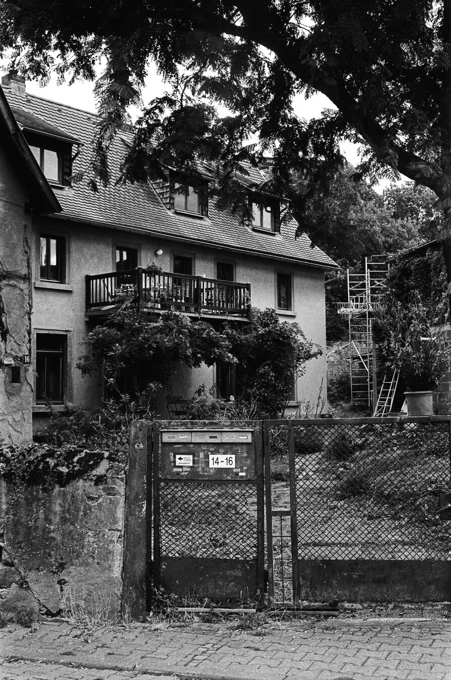 Another, larger residential building on the same property. Shrubs that reach up to the balcony on the first floor surround the entrance area. The trunk of a large tree juts into the photo from the right. In the foreground, a fence with an entrance gate can be seen. The corner of the house from the previous photo can still be seen on the left edge of the picture.