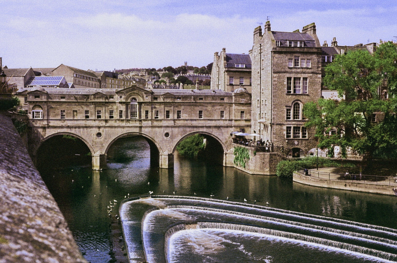 Analog color photo of the Pulteney Bridge in Bath, England. The iconic bridge, with its three arches standing in the water, houses several cafés and shops whose windows are visible along the span. The elliptical structures in the shallow water create little waterfalls, numerous seagulls enjoying life there. The houses of Bath are visible behind the bridge against a blue sky with light, wispy clouds. The colors in image number 1 have been edited.
