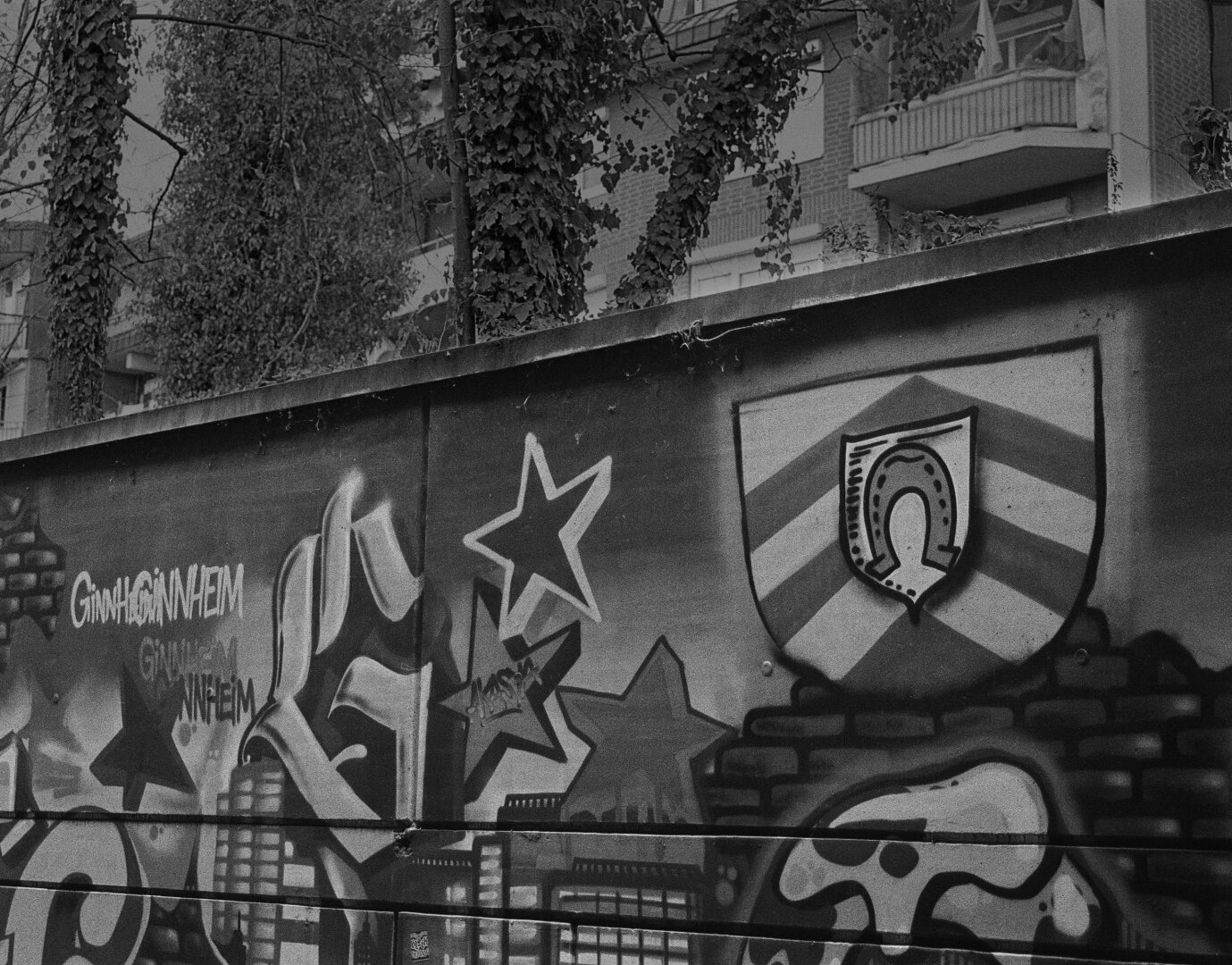 Analog black snd white photo of a wall with Ginnheim Graffitti. Also the coat of arms of Ginnheim, a horseshoe with stripes in the background, is sprayed there. Behind the wall windows and balconies of an apartment house, build in the late 80s, can be seen.