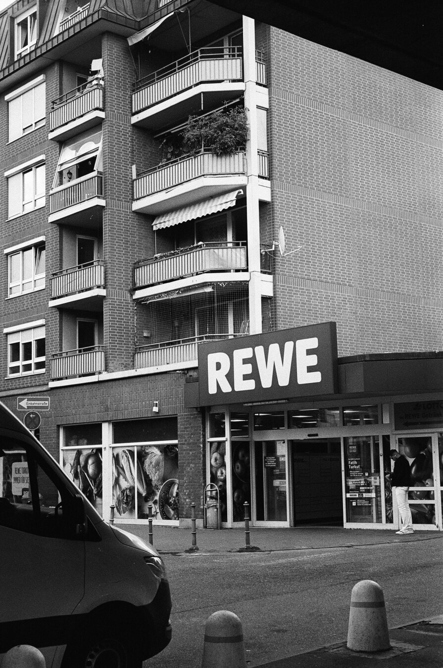 Brutalism at its best: the corner of a huge apartment house (the same as shown in the shot before). On the ground level is an entrance to a supermarket. A huge „Rewe“ sign is above the entrance door.