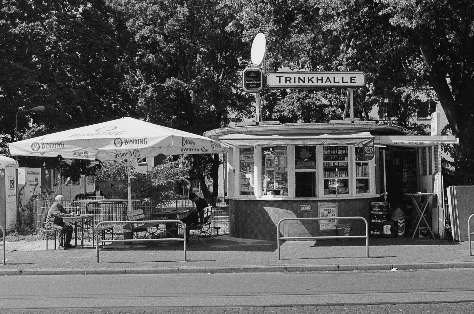 The same subject as before, but with the camera held horizontally. Now a large umbrella can be seen to the left of the kiosk, under which there are benches. Two men sitting there, enjoying their cold brew.