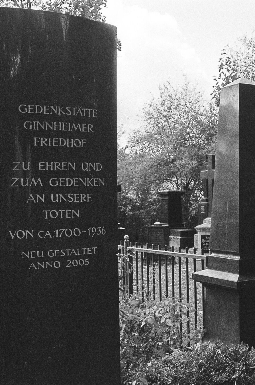 Memorial stone with the inscription: „Ginnheimer cemetery memorial in honor and remembrance of our dead. From about 1700 - 1936. Redesigned in 2005“. Graveyards and gravestones in the background.