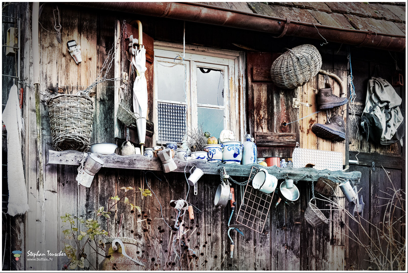 Verwitterte Holzhütte mit Fenster, davor ein Regal voller Gegenstände: Emailletassen, blau-weiße Keramikkrüge, Moka-Kanne, Weidenkörbe, Regenschirm, Wanderschuhe, Rucksack und diverses Küchengeschirr. Kupferne Dachrinne, rankende Pflanzen.