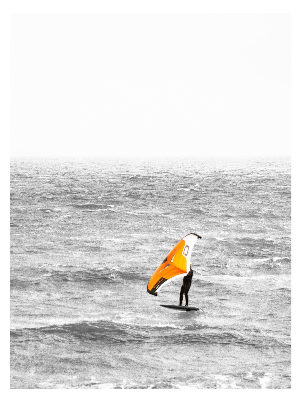 Das Bild zeigt einen Kiter mit einem orangen Kite vor Küste in East Sussex. Das Meer ist vom starken Wind aufgewühlt. Der Himmel ist grau und in der Ferne scheinen Himmel und Meer zu verschmelzen
