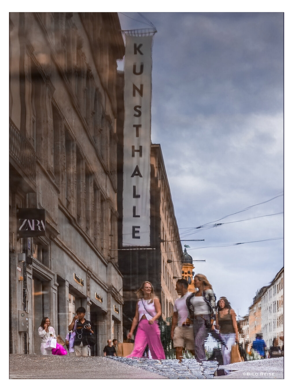 Eine Gruppe von Menschen spiegelt sich in einer großen Pfütze. In dem Bild ist ausschließlich die Spiegelung zu sehen, diese ist aber horizontal gespiegelt. Den Hintergrund bildet eine Einkaufsstraße in München.