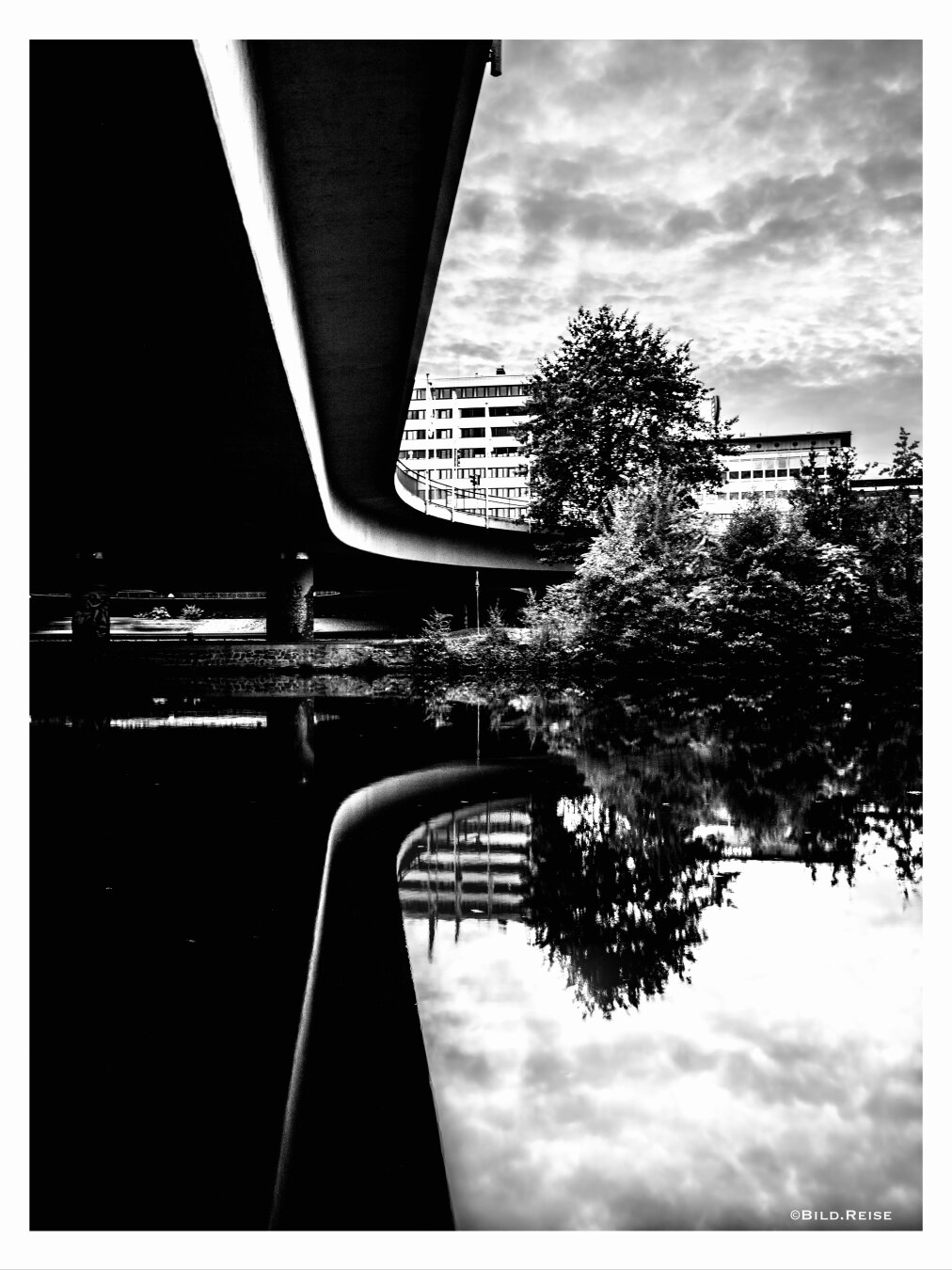 Schwarzweißfoto einer Brücke von unten (linke Bildhälfte) die Brücke spiegelt sich im Fluß. Auf der rechten Bildhälfte sind Hochhäuser und Bäume zu sehen, die sich ebenfalls im Fluß spiegeln.