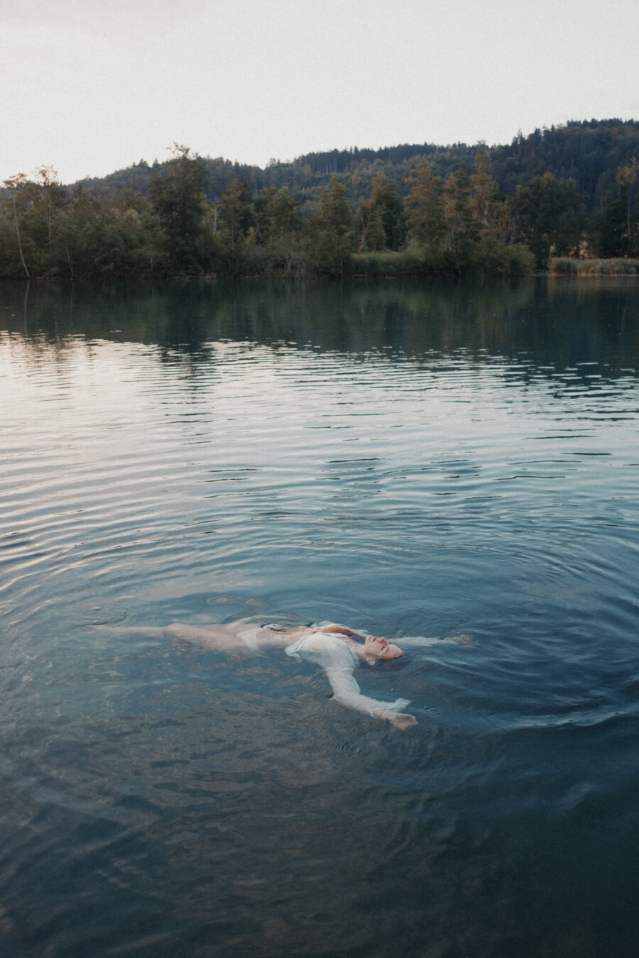 A person floats peacefully on their back in calm blue lake water, arms spread wide. They wear a flowing white garment that drifts on the water's surface. Gentle ripples surround them. Behind, dark forested hills meet a soft gray sky along the shoreline.