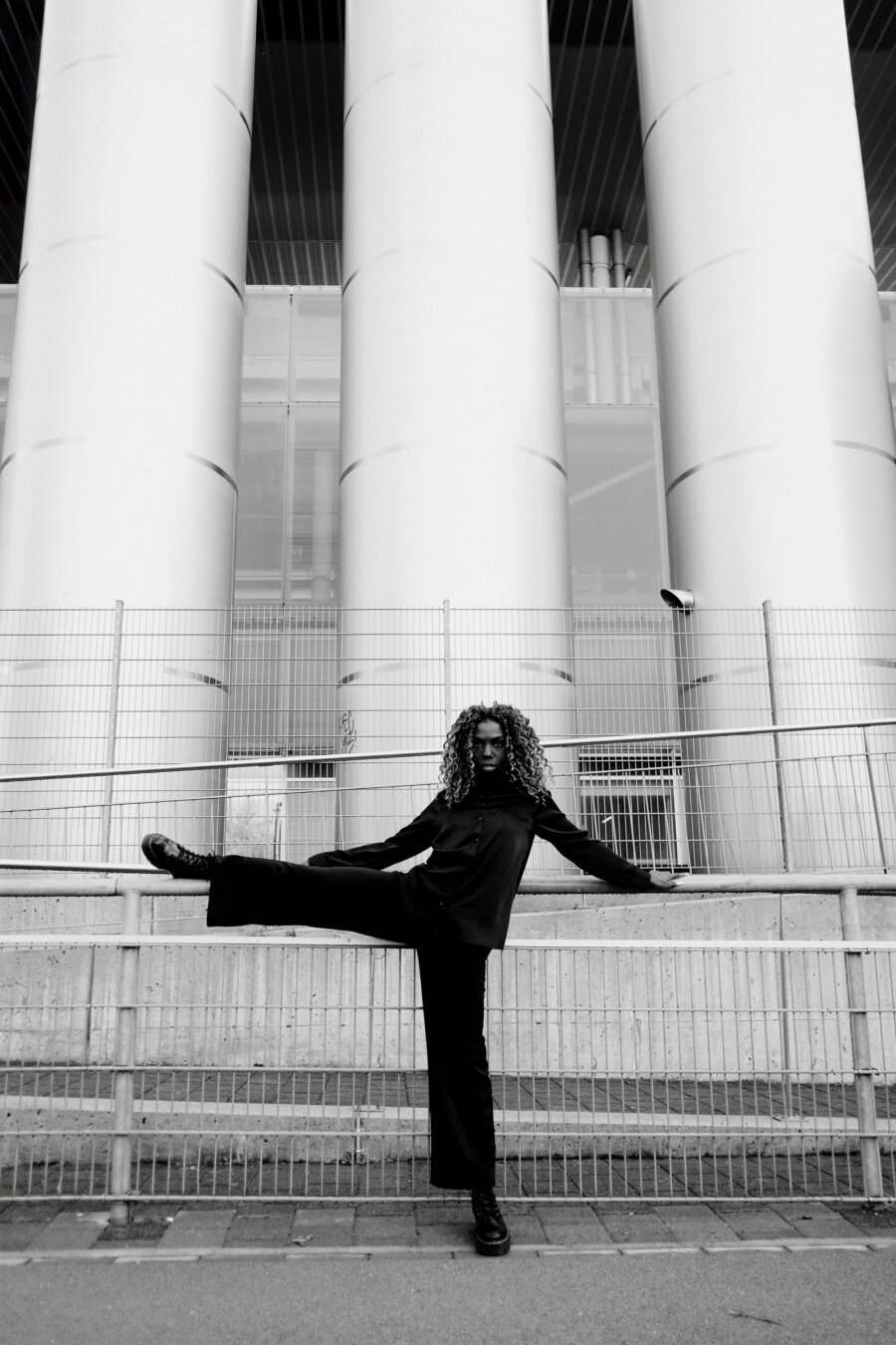 A black-and-white full-body shot of a person with voluminous curly hair performing an athletic pose, one leg extended and resting on a metal railing. They wear all-black clothing and look directly at the camera. Three massive cylindrical columns and a modern building facade dominate the architectural background behind wire fencing.