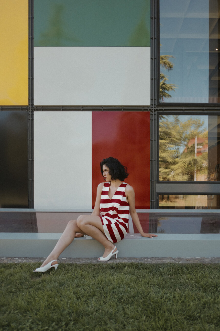 A woman in a red-and-white striped dress and white heels sits on a concrete ledge in front of a colorful modern building with geometric panels in yellow, green, white, and red. She gazes away from the camera toward the left side. Green grass in the foreground; tree reflections visible in windows to the right.