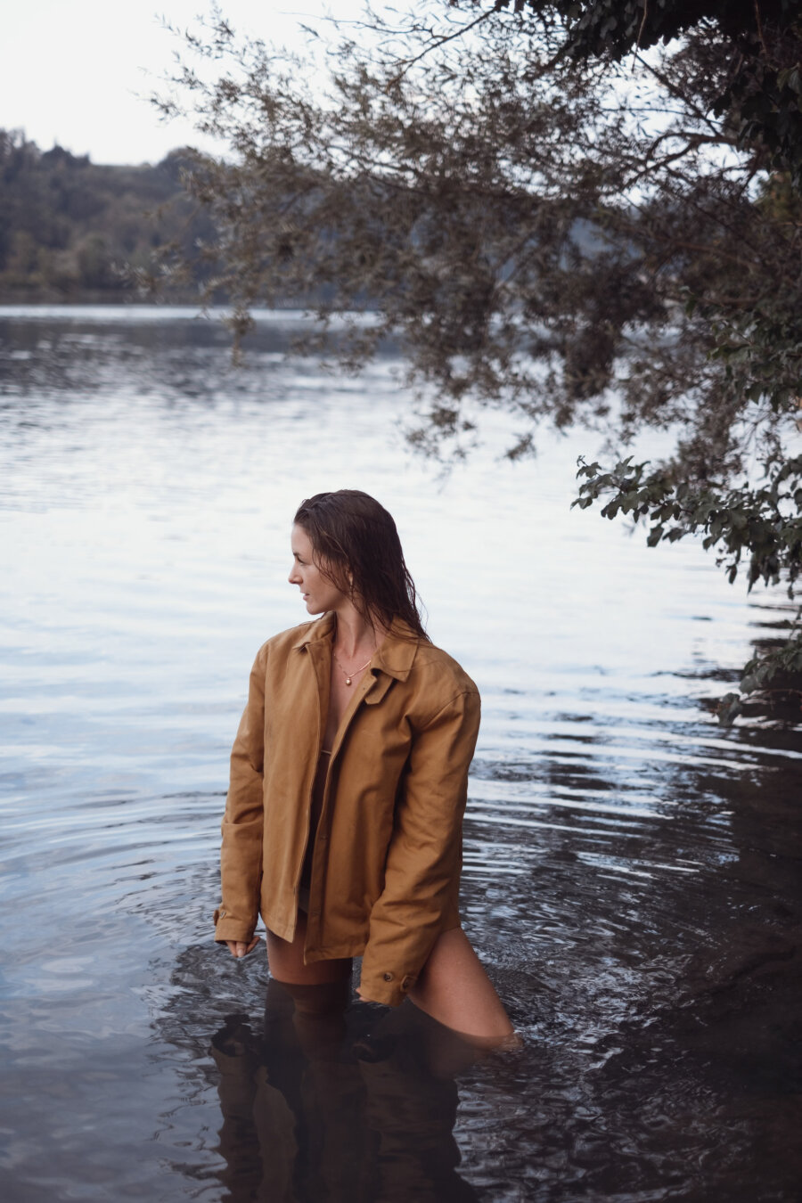 A woman with wet hair stands in a calm lake wearing an open brown jacket, looking toward the shore with tree branches hanging above her.