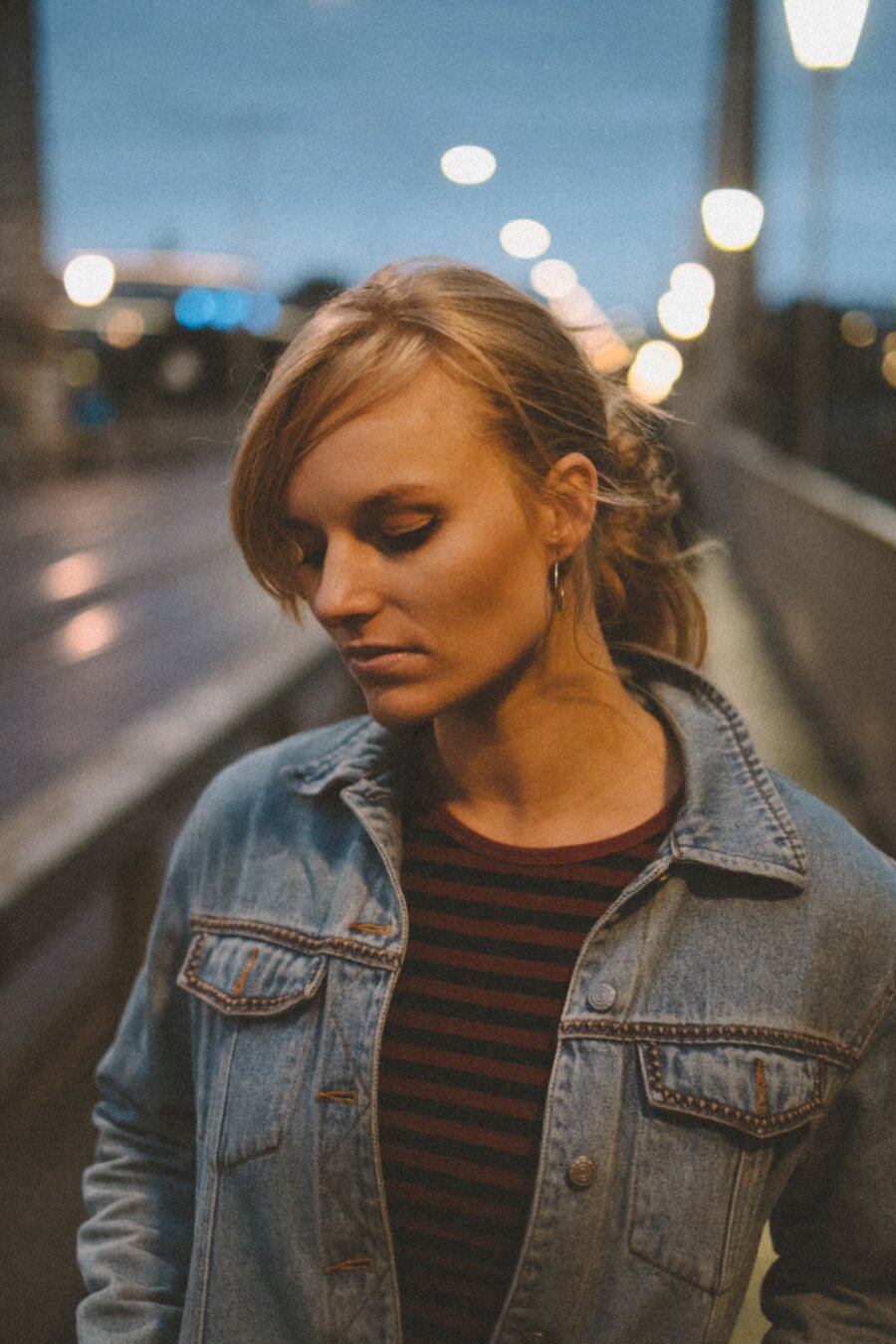 A young woman with blonde hair tied back stands outdoors at dusk, looking downward in a contemplative pose. She wears a denim jacket over a red-and-black striped shirt and small hoop earrings. Soft bokeh city lights glow in the blurred background.