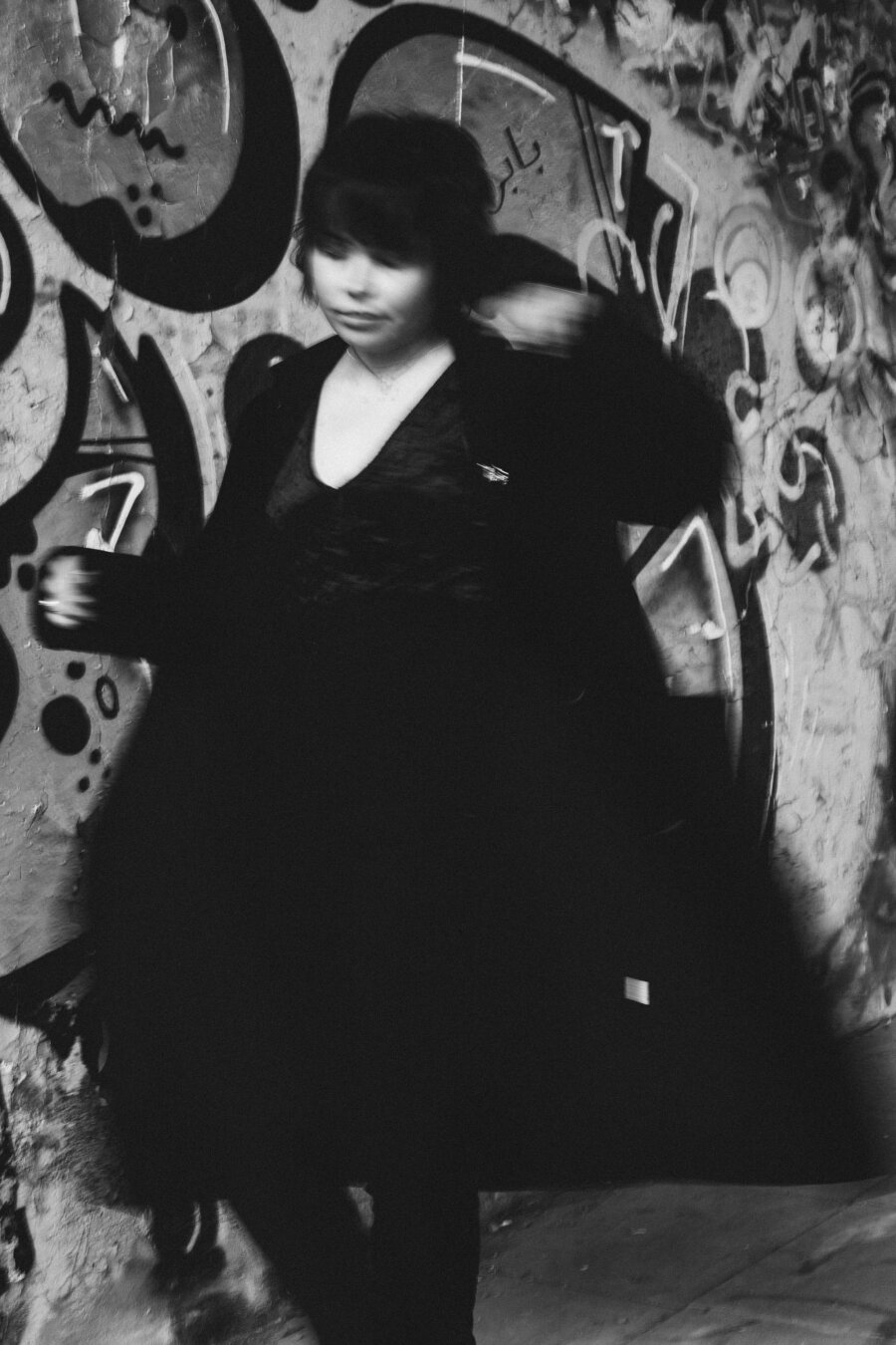 A black-and-white portrait of a person with dark hair standing against a graffiti-covered wall. They wear dark clothing and appear captured mid-motion with slight blur on one arm. Abstract graffiti designs with curved shapes and text elements fill the textured background.