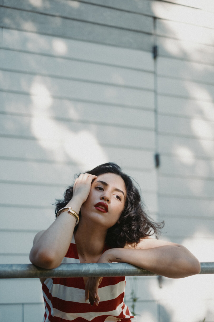 A woman with dark wavy hair and red lipstick leans on a metal railing against a white slatted wall. She wears a red-and-white striped top and a gold bracelet, with her hand gently resting by her face as she looks upward. Natural sunlight casts tree shadows on the wall behind her.