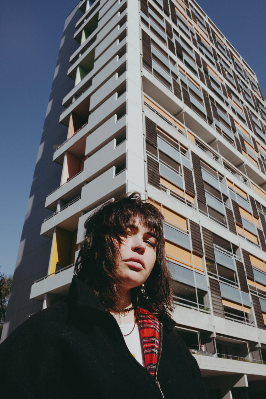 A person with dark shaggy hair and bangs stands in front of a modern apartment building with yellow and gray balconies. They wear a black coat with a red plaid scarf. Shot from below against a clear blue sky.