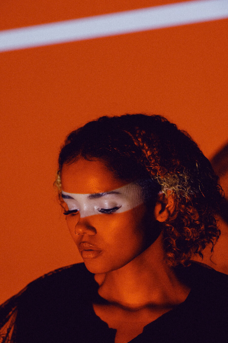 Portrait of a person with dark skin and curly hair shown in profile with downcast eyes, bathed in warm orange light. A white horizontal stripe crosses their forehead above the eyes. Black textured top against a solid orange background. Contemplative and serene mood.