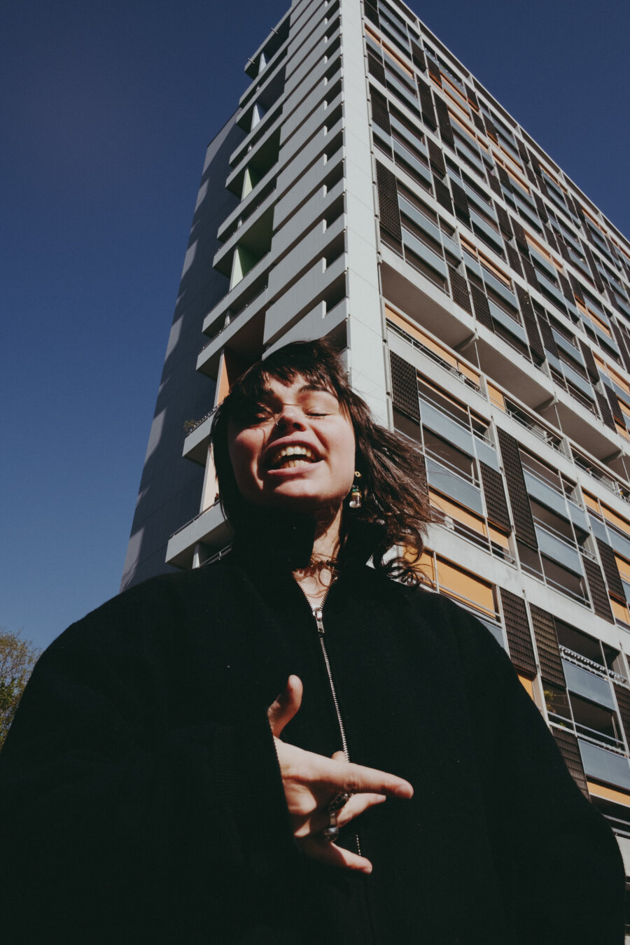 The person laughs with eyes closed in front of the same apartment building. Their hand gestures forward, wearing a black zippered jacket and rings. Dynamic low-angle shot capturing motion and expression.