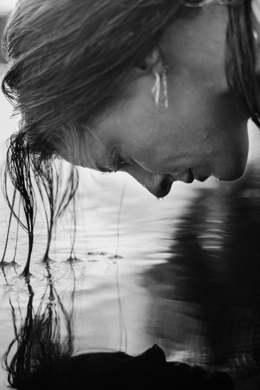 Black-and-white close-up portrait of a person with long wet hair leaning over rippling water, head bowed in profile, face nearly touching the surface, soft reflections stretching across the water.