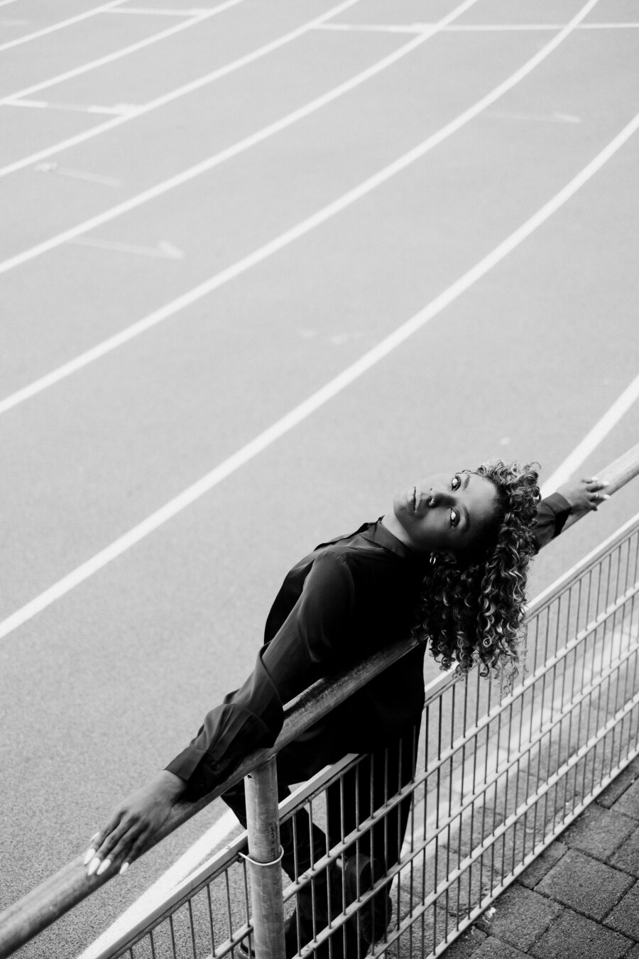 Black-and-white photo taken from above: a Black woman with long curly hair leans back over a metal railing, arms stretched out along the handrail, looking up into the camera, a running track with white lane markings stretching out below her.