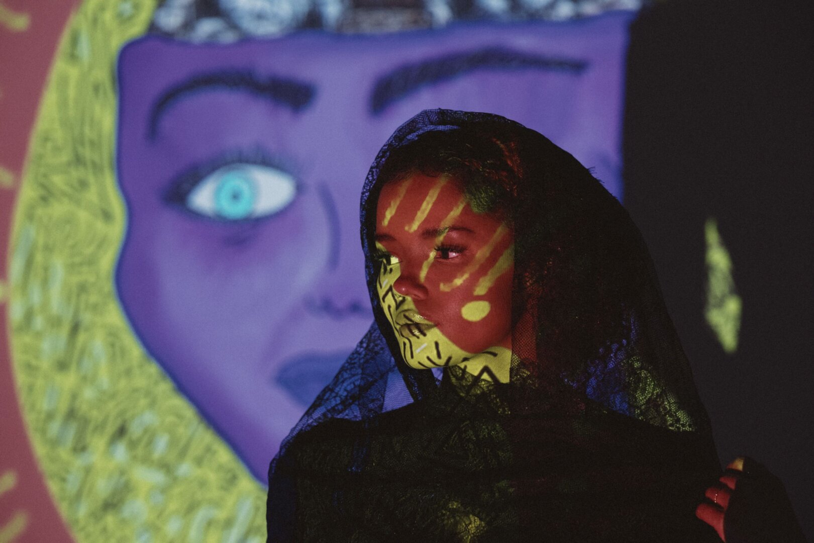 Person wearing black lace veil with yellow geometric light patterns projected onto their face. Behind them, a large purple-toned projection shows a stylized face with a striking blue eye.
