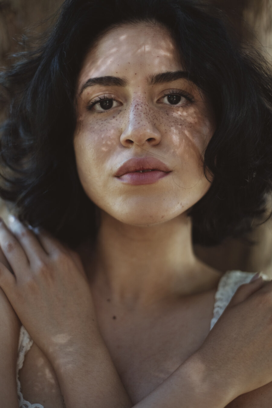 Close-up portrait of a person with shoulder-length dark curly hair and visible freckles across their nose and cheeks. They're wearing a white lace-trimmed top, with their hand resting near their shoulder. Soft natural lighting creates gentle shadows on their face.