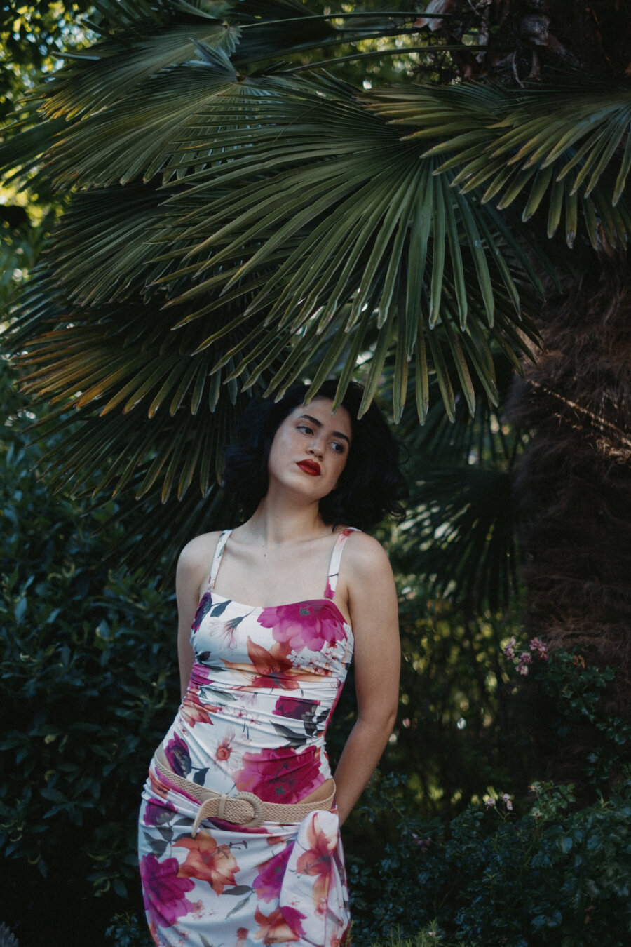Woman with dark wavy hair and red lipstick stands beneath a large palm frond, wearing a white floral dress in pink and orange tones with a woven belt, surrounded by lush green foliage in soft evening light.