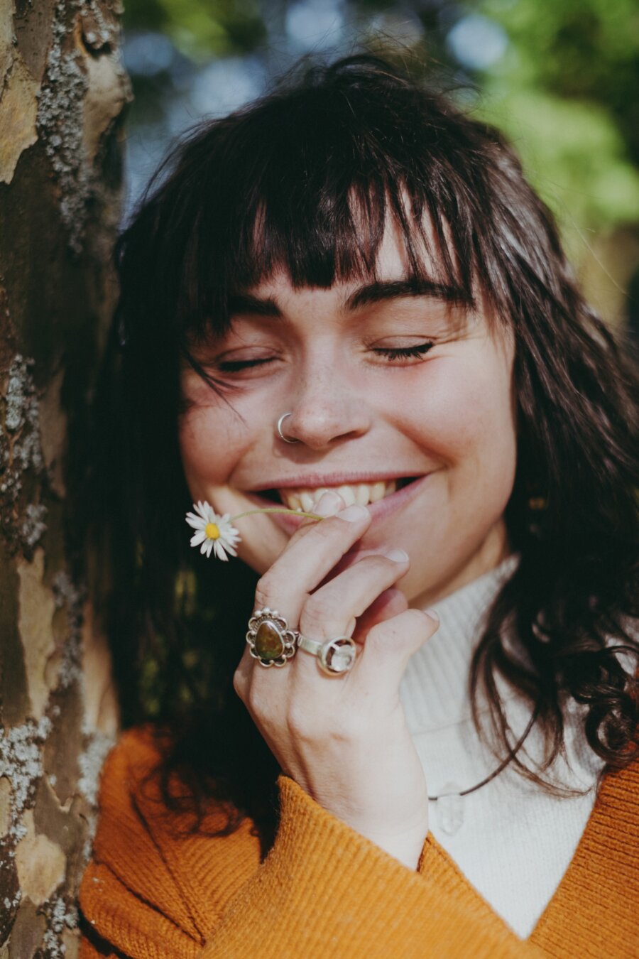 Person with dark wavy hair and bangs smiling with closed eyes, holding a small white daisy near their mouth. They have a silver nose ring and multiple rings on their fingers, wearing an orange cardigan over a white top, standing beside a tree trunk in natural daylight.
