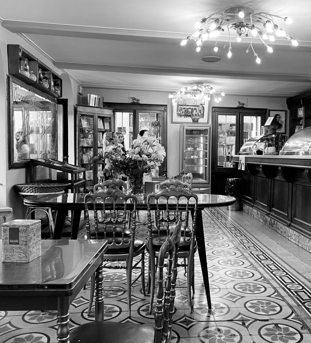 Schwarz-weiß Aufnahme eines alten, traditionellen Cafés und Bäckerei in einer kleinen Ortschaft an der Ligurischen Küste, Italien.
Tische, Stühle, andere Einrichtungsgegenstände und das Muster der Bodenfliesen erinnern an eine vergangene Zeit. Alles ist aber sehr gut gepflegt. Die ruhige und freundliche Atmosphäre laden hier zu einer Pause mit Cappuccino und ein Törtchen ein.

A black-and-white photograph of an old, traditional café and bakery in a small village on the Ligurian coast, Italy.
The tables, chairs, other furnishings, and the pattern of the floor tiles evoke a bygone era, yet everything is very well maintained. The quiet, welcoming atmosphere invites visitors to pause and enjoy a cappuccino and a small pastry.