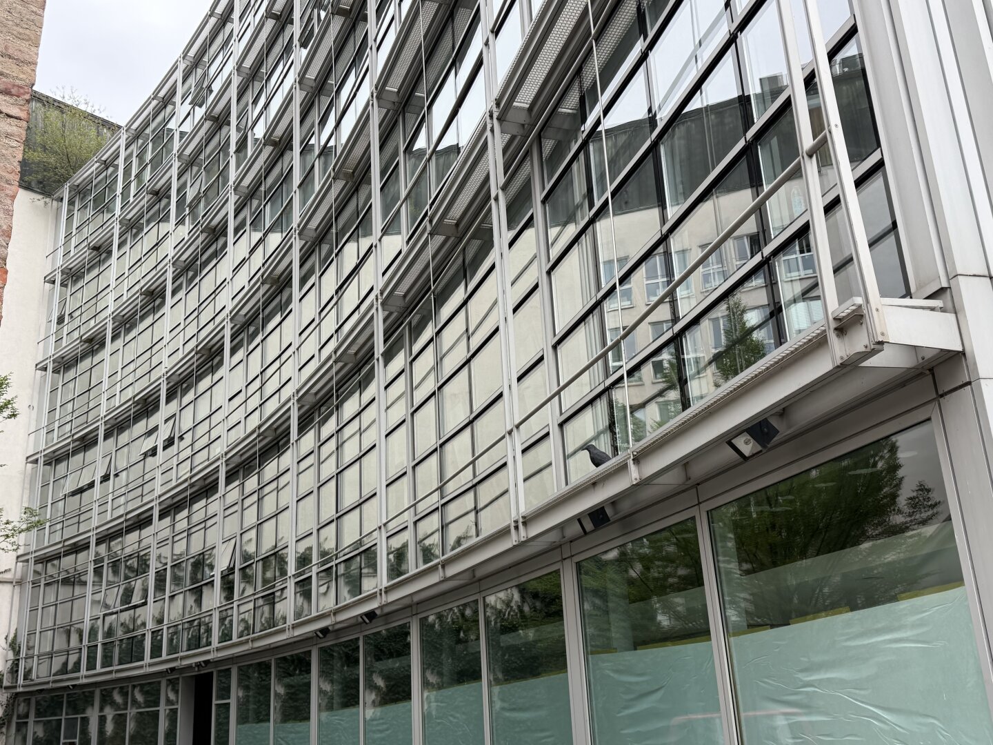 A modern glass and steel building facade in Munich. It appears to be an office and commercial building. The facade curves inward toward the building's interior. The surroundings—walls, older buildings, and trees—are reflected in the large glass surfaces.