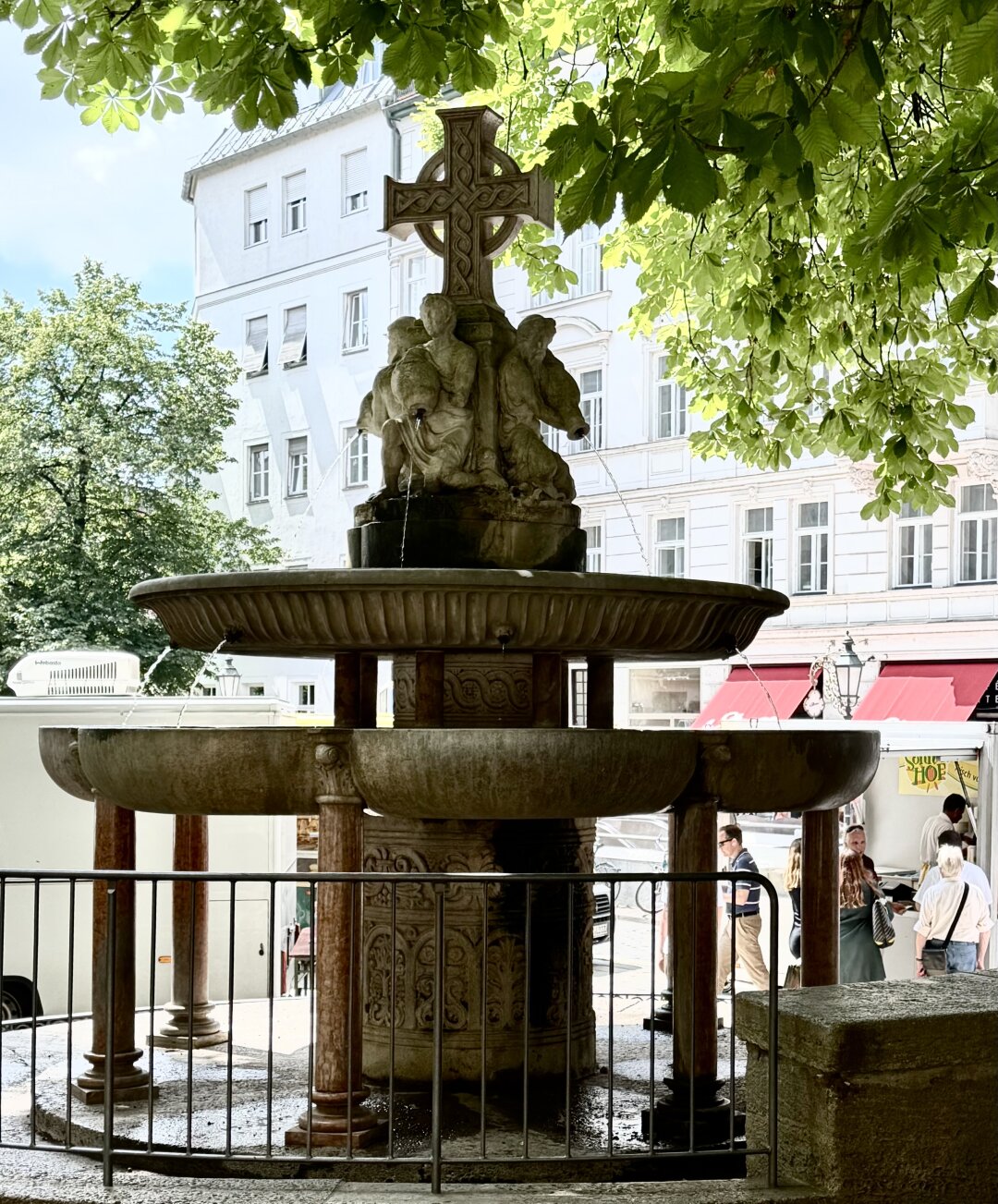 Hier: Blick im Gegenlicht vom Kirchenvorplatz in Richtung Markt.

St. Anna Fountain (tiered-basin fountain), created in 1894 by Anton Pruska based on a design by Gabriel von Seidl; located at the corner of the terrace southwest of the church.  View from the church forecourt towards the market.