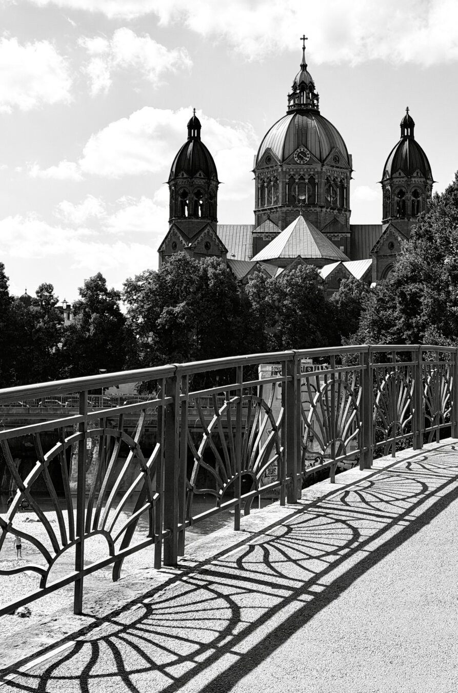 A view of St. Luke's Church. The church is located by the Isar River, and the black-and-white photo was taken from a bridge spanning the river. The bridge railing is visible in the foreground, while the upper part of the church can be seen on the other side of the Isar, behind the trees.