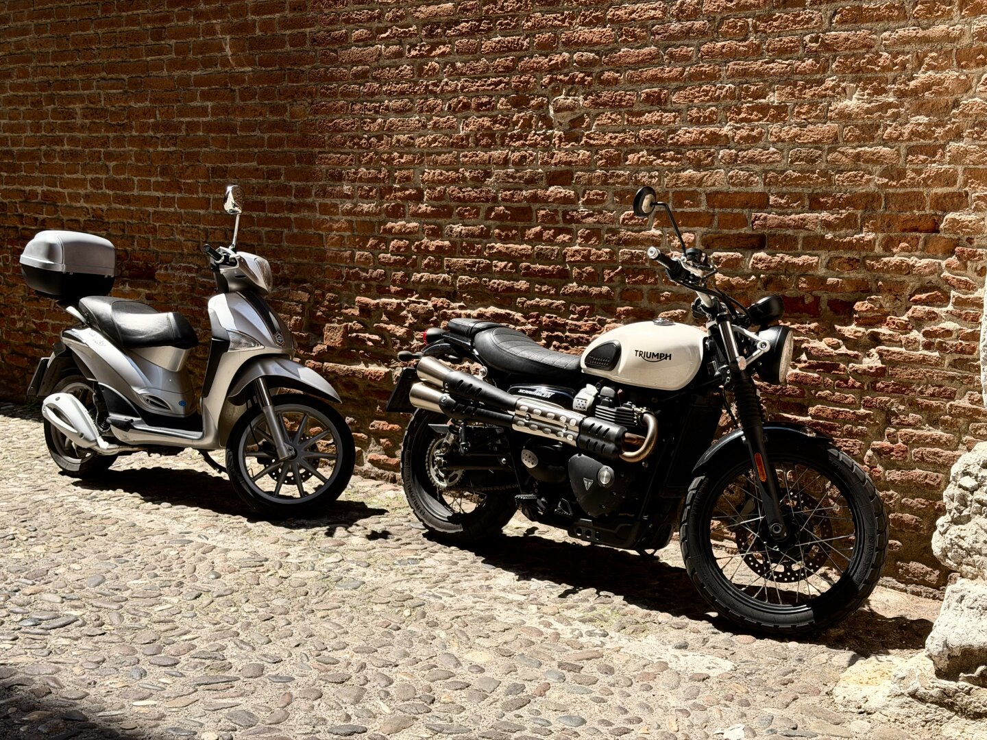 Two motorcycles are parked in the midday heat in a narrow street, within a white marked area. They are positioned right in front of  a side wall of the Cathedral of Sant’Anastasia in Verona. The wall behind them is made of brick. The street surface consists of beige-gray cobblestones.