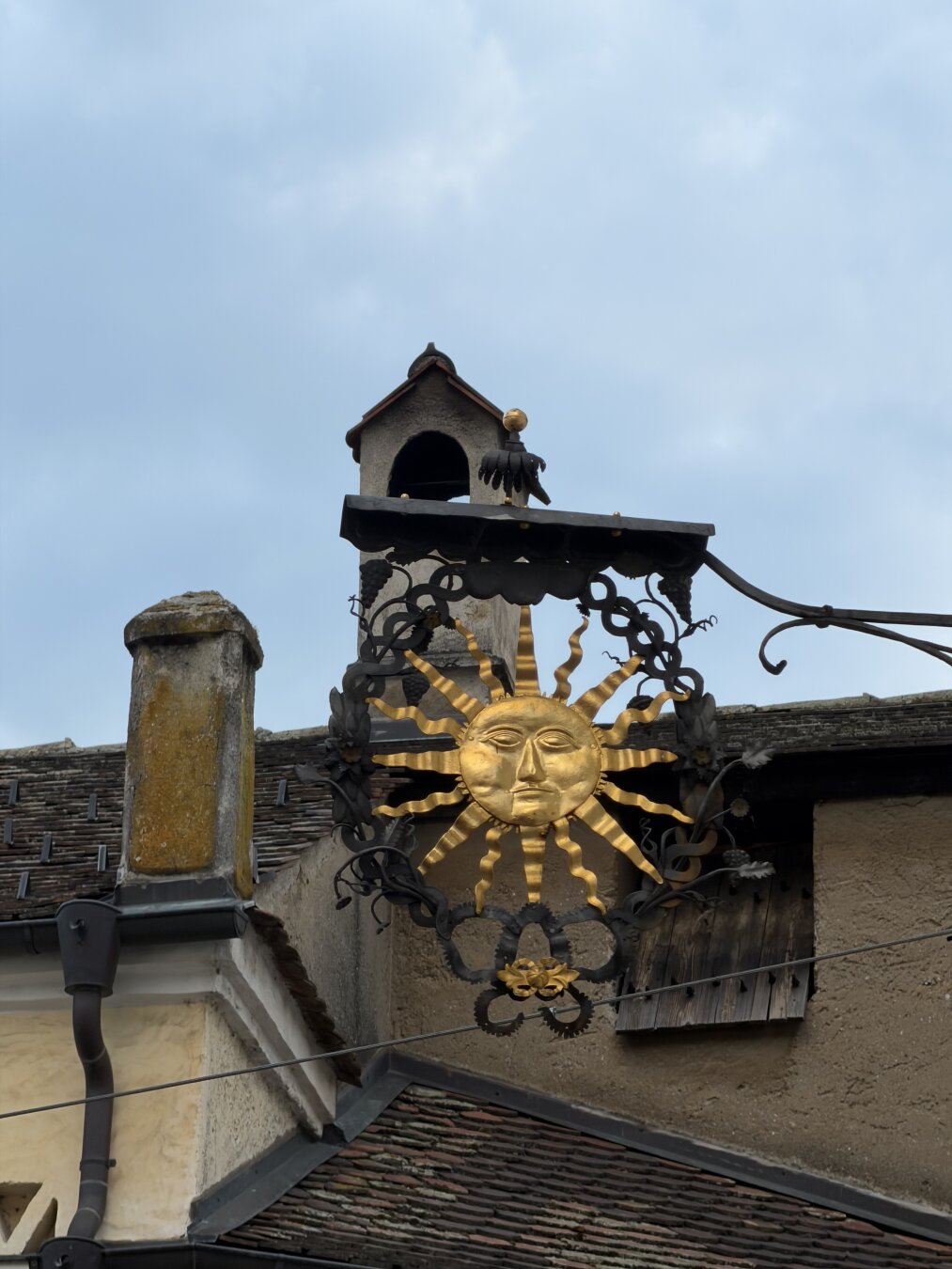 Im Mittelpunkt des Bildes sieht man ein Schild, das eine Sonne darstellt.
Die Sonne hat eine  goldenen Farbe und ist mit einem Fries aus schwarzem Schmiedeeisen eingerahmt. Im Hintergrund sind Dächer mit Ziegeln und einen Himmel mit sehr leichten Wolken im Abendlicht zu sehen.

At the center of the image, one sees a sign depicting a sun.
The sun is golden in color and framed by a black wrought-iron border. In the background, tiled roofs and a sky with wispy clouds in the evening light are visible.