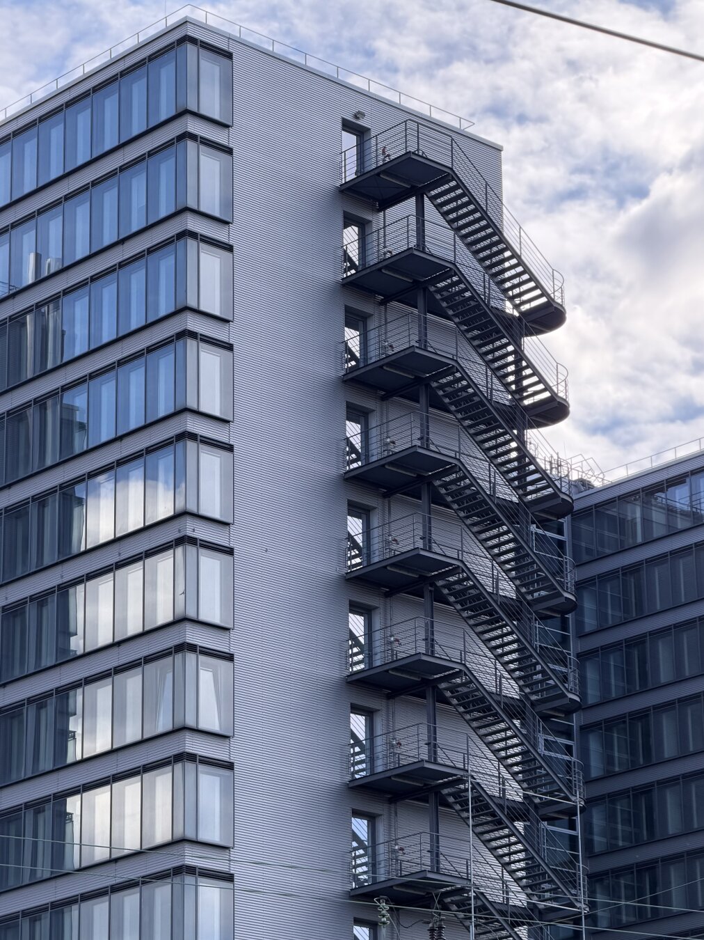 Monochrome details of two high-rise buildings. Set against a pale blue sky with white clouds, two interconnected high-rises are visible; both buildings feature facades with large expanses of glass. The surroundings are reflected in the large windows. The afternoon sun creates bold highlights and shadows, even on the lower sections of the buildings. The facade visible in the center of the image has less window area; darker steel staircases with landings leading to the glass doors stand out prominently here. The steps of the external staircase and the cloudy sky are reflected in the glass doors that open onto the landings on each floor. The photo was taken from an S-Bahn station in Munich