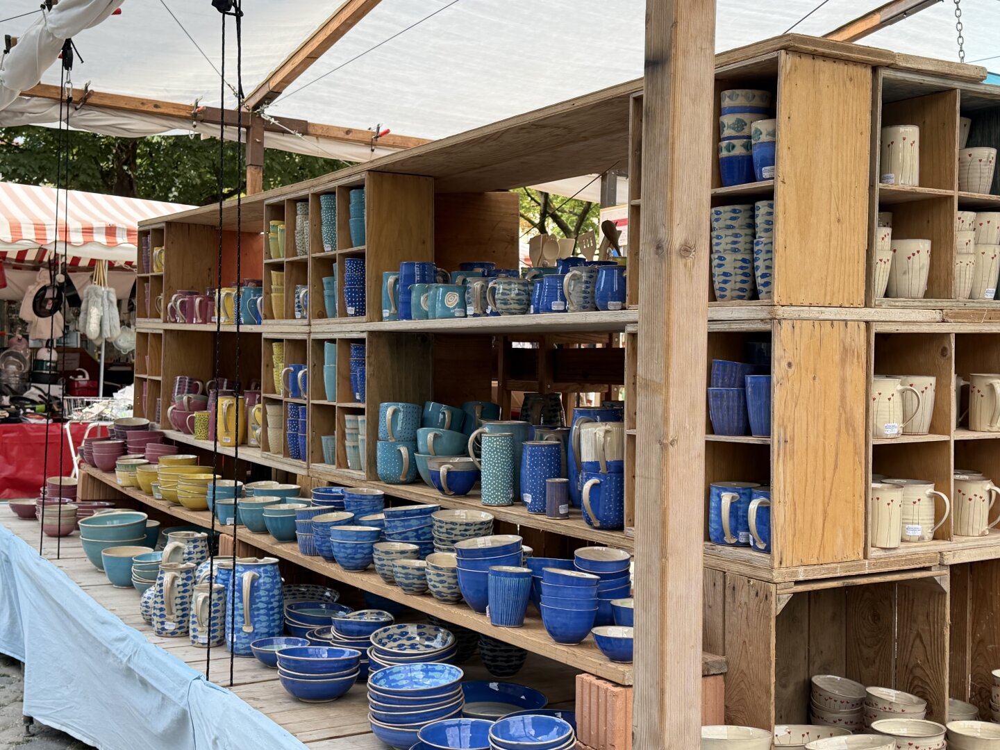 Two wooden stalls can be seen at the Auer Dult in Munich. On the shelves of the stalls, one can see many ceramic household items in various colors and patterns.