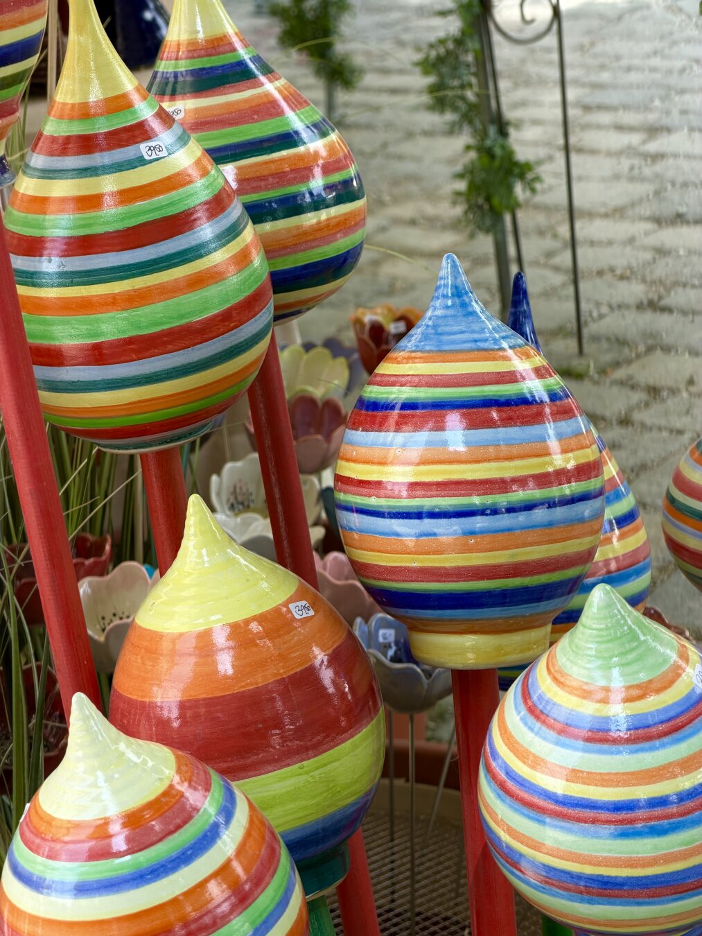 A close-up of colorful ceramic garden spheres. These spheres are painted in bold colors and feature stripes of varying widths. They are mounted on red rods. The photo was taken at a stall selling pottery garden art at the Auerdult market in Munich.