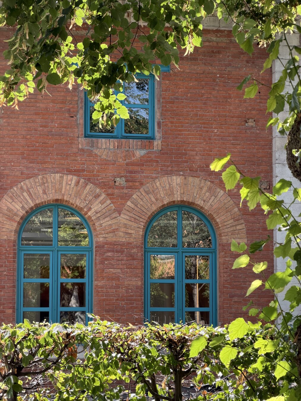 Detail of the brick façade of the former locomotive shed (now an exhibition center) in Rosenheim. A rectangular window is visible on the upper floor, along with round-arched French windows on the ground floor. The window frames are turquoise-blue, and the surrounding trees are reflected in the large windowpanes.