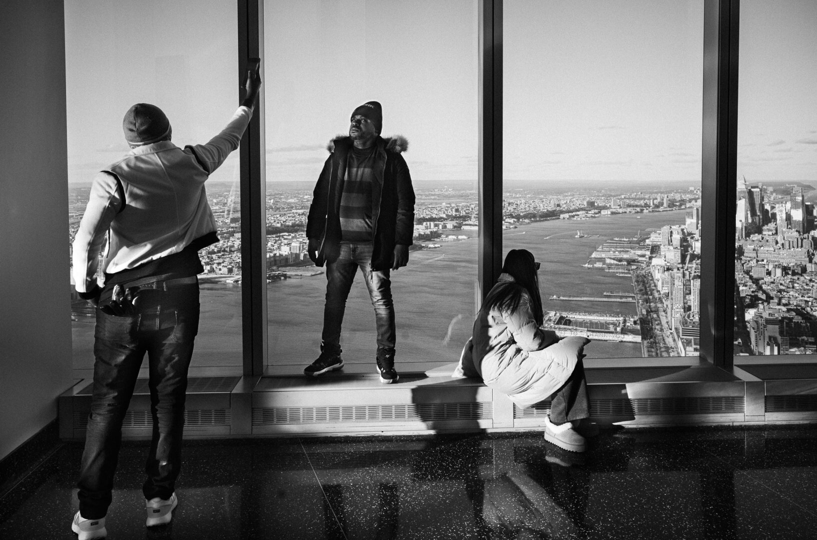 Black-and-white street photograph of three people beside the large windows of a New York observation deck, each facing or using the view differently, with strong sunlight and reflections across the floor.